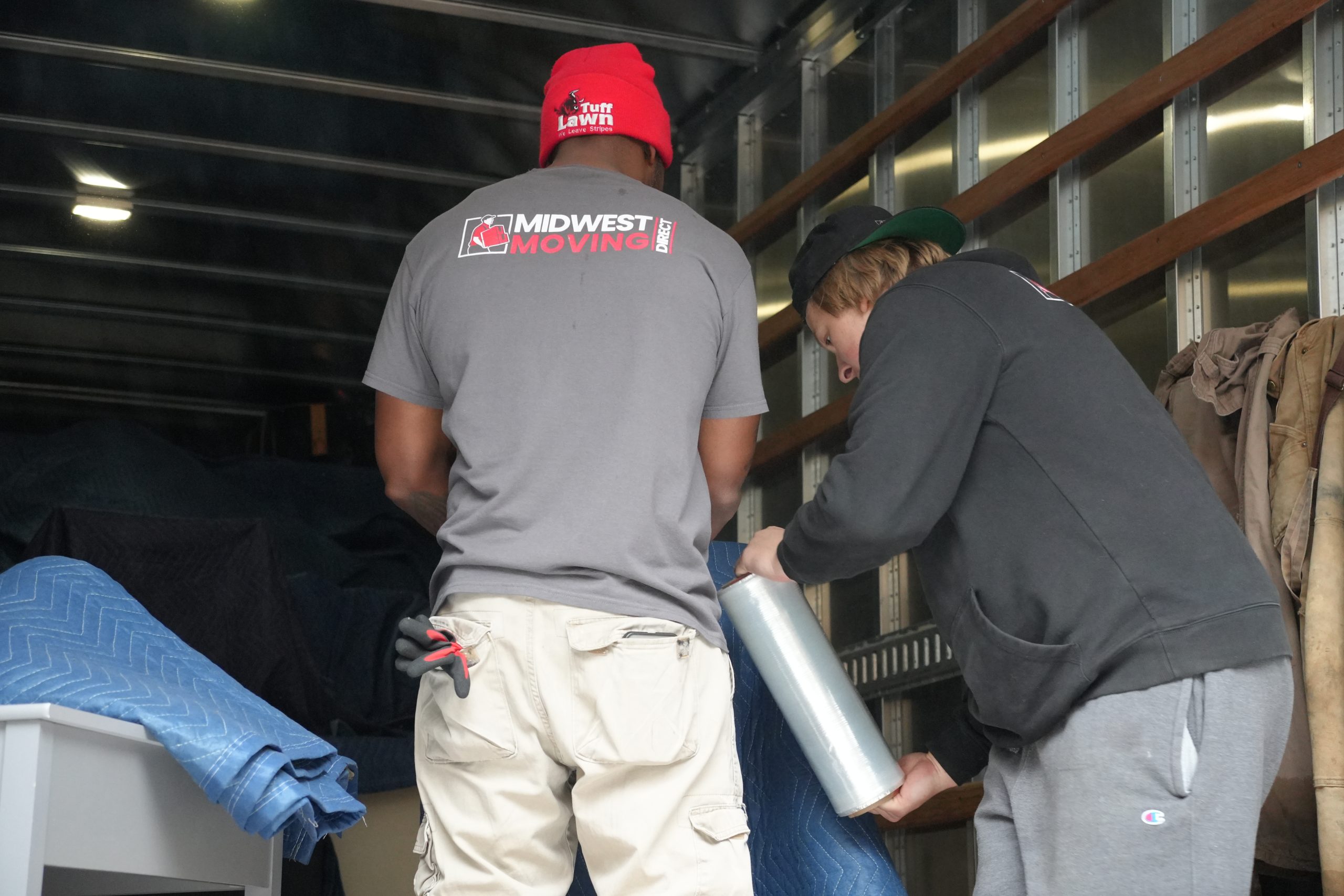 Two Midwest Moving Direct employees preparing for a move, one wrapping furniture with stretch film and the other organizing moving blankets inside a truck.