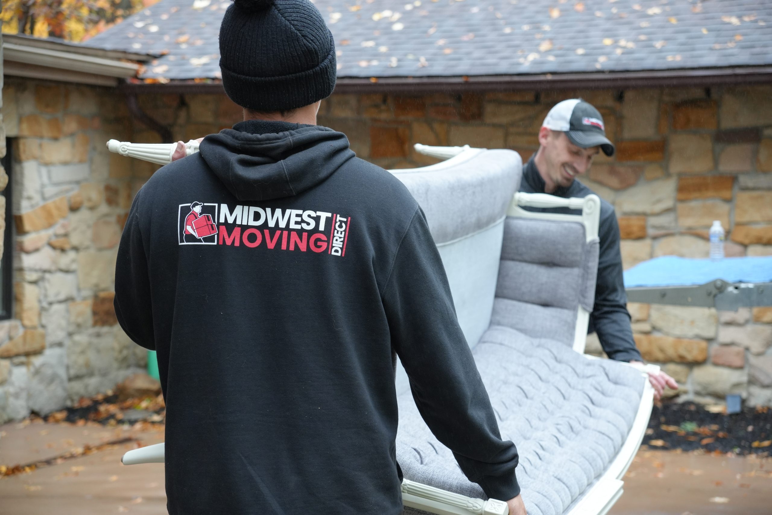 Two Midwest Moving Direct movers carrying a gray upholstered sofa outdoors, showcasing professional relocation services in Akron, Ohio.