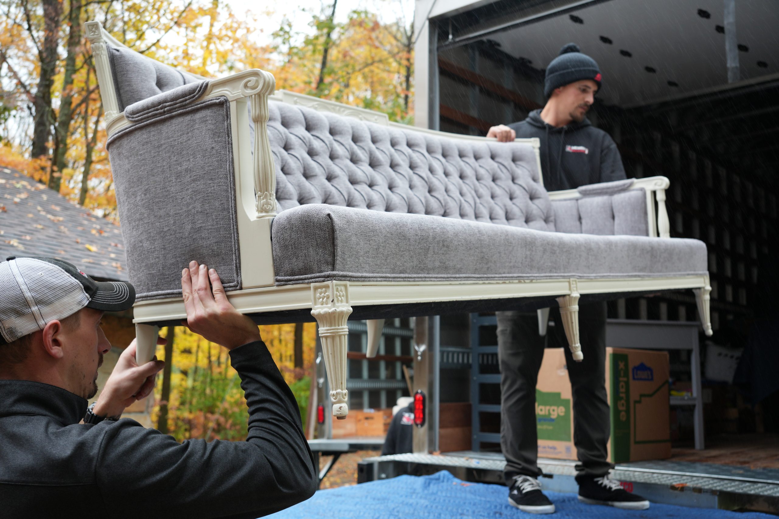 Two movers carefully lifting a gray upholstered sofa with ornate white legs onto a moving truck, surrounded by autumn foliage, illustrating professional moving services.