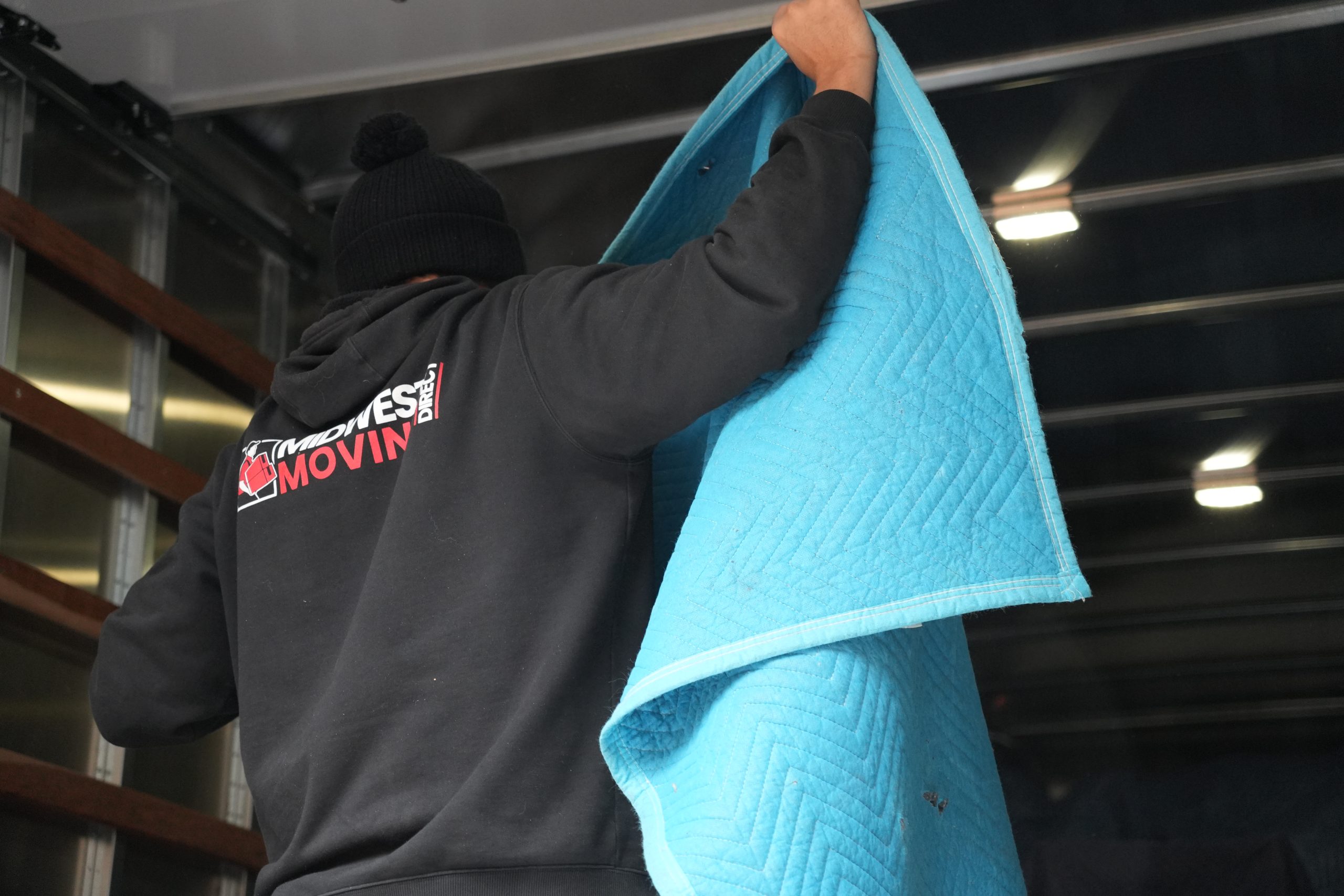 Mover in black hoodie with "Midwest Moving Direct" logo, lifting a blue moving blanket inside a truck.